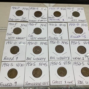 May include: A collection of vintage wheat pennies, each encased in a small white cardboard holder. The holders are labeled with dates, grades, and notes about the coins, such as "CUD LEFT WHEAT" and "BIE LIBERTY".