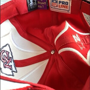 May include: Red and white baseball cap with a white NFL Pro Line logo and a red and white Kansas City Chiefs logo. The cap has a white adjustable strap with a black buckle.