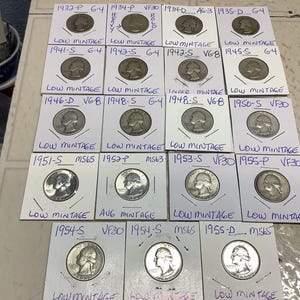 May include: A collection of silver quarter coins, each encased in a small white cardboard holder. The holders are labeled with dates, mint marks, and grading information, such as "Low Mintage" and "VF30". The coins are arranged on a light-colored surface.