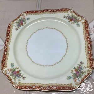 May include: Square cream-colored plate with a floral design and a decorative red border. The plate features a central white area and is trimmed with gold accents. The corners have a scalloped edge.