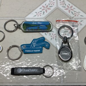 May include: A collection of seven keychains with various designs. The keychains include a blue and white keychain with a picture of Lake Tahoe, a blue keychain with the words "Peerless Faucet", a silver keychain with the word "Lowe's", a white keychain with the words "Ace Hardware", a black keychain with the words "OmniSource Corporation", and two silver keychains with no designs.