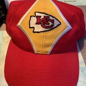 May include: Red and yellow baseball cap with a white and red Kansas City Chiefs logo. The logo is a stylized arrowhead with the letters "KC" inside.