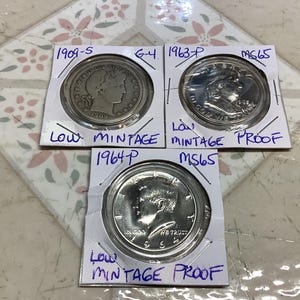 Può includere: Tre monete d'epoca in supporti protettivi. Le monete sono datate 1909-S, 1963-P e 1964-P. Ogni supporto è etichettato con testo scritto a mano, tra cui "Low Mintage Proof" e "MS65". Le monete sono di colore argento.