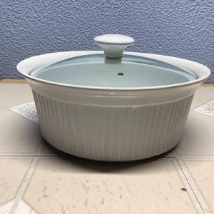May include: White ceramic casserole dish with a glass lid. The dish has a ribbed exterior and a white knob on the lid. The dish is sitting on a tiled surface.