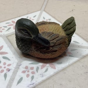 May include: A detailed figurine of a duck, featuring a dark green head and a black beak. The duck's body is a mix of brown and tan, with textured feather details. The duck is resting on a patterned surface.
