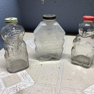 May include: Three vintage glass coin banks. One is shaped like an elephant with a green lid, another is a geometric shape with a black lid, and the third is a bear with a red lid. All are empty.