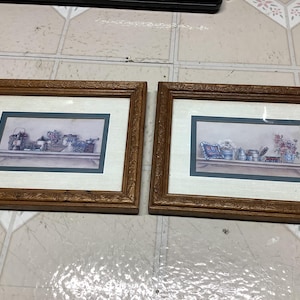 May include: Two framed prints with a vintage aesthetic. Each print features a shelf with various decorative items, including vases, teacups, and other collectibles. The frames are a warm brown color, and the prints have a soft, muted color palette.