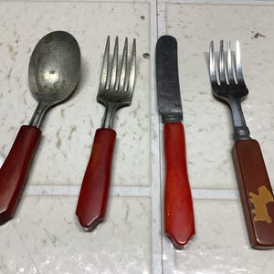 May include: A set of vintage silverware with red and brown handles. The set includes a spoon, two forks, and a knife. One fork has a brown handle with a gold-colored dog silhouette. The silverware is arranged on a light-colored surface.