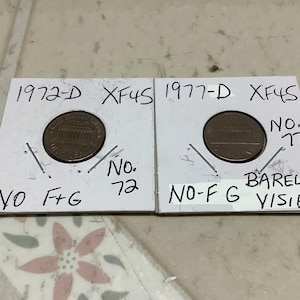 May include: Two coin holders with pennies. The left holder has a penny from 1972-D, marked XF45, with the text "NO F+G" and "No. 72". The right holder has a penny from 1977-D, marked XF45, with the text "NO-F G" and "BARELY VISIBLE".