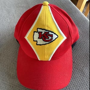 May include: Red baseball cap with a yellow diamond-shaped patch featuring the Kansas City Chiefs logo. The logo is embroidered in white and black with the letters "KC" and an arrowhead.