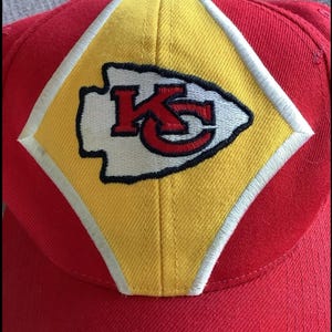 May include: Red and yellow baseball cap with a white and red embroidered Kansas City Chiefs logo. The logo features the letters "KC" inside an arrowhead.