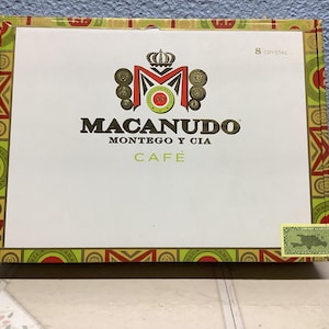 May include: A rectangular box with the brand name "MACANUDO" in brown lettering. The box features a decorative border with green, red, and yellow patterns. The text "MONTEGO Y CIA CAFÉ" is also visible.