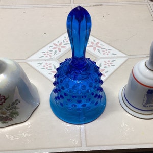 May include: Three decorative bells, one blue glass bell with a textured surface, one white porcelain bell with a floral design, and one white porcelain bell with a red, white, and blue design and the text "Six Flags".
