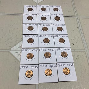 May include: A collection of 12 copper-colored US coins from 1956 to 1958, displayed in individual white card holders. Each coin is labeled with the year, mint mark, and grade.