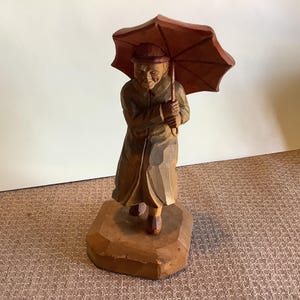 May include: A wooden carving of a person walking in the rain, holding an umbrella. The figure is wearing a long coat and hat, and the umbrella is open. The carving is mounted on a wooden base.