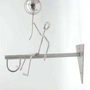 Könnte beinhalten: Silberfarbene Metallskulptur eines Strichmännchens, das eine Kugel hält und auf einer horizontalen Stange zu sitzen scheint. Die Stange ist mit einer dreieckigen Halterung an der Wand befestigt. Die Skulptur hat ein modernes, minimalistisches Design.
