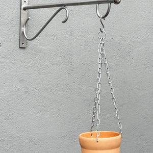 Könnte beinhalten: Eine silberfarbene Wandhalterung aus Metall mit einem horizontalen Arm und einer S-förmigen Stütze. Ein terrakottafarbener Blumentopf hängt an der Halterung an einer silbernen Metallkette, die mit einem S-Haken am Arm befestigt ist.