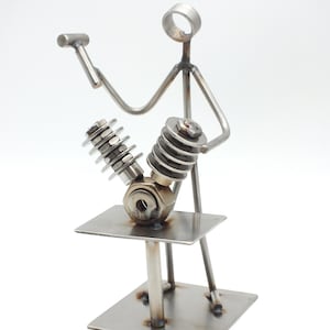 Könnte beinhalten: Eine Metallskulptur einer Figur, die an einem Motor arbeitet. Die Figur besteht aus Metallstäben und hält einen Hammer. Der Motor besteht aus verschiedenen Metallteilen, darunter Federn und Zylinder. Die Skulptur ist auf einem quadratischen Sockel montiert.