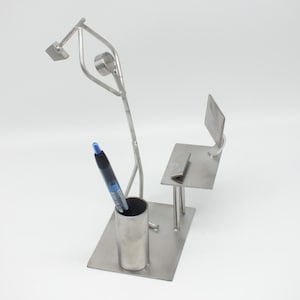 Könnte beinhalten: Eine Schreibtisch-Organizer-Skulptur aus gebürstetem Metall. Die Skulptur umfasst einen Schreibtisch, einen Stuhl und eine Lampe mit einem Stift in einem zylindrischen Halter. Der Schreibtisch ist rechteckig, mit einem kleinen Stuhl und einer Lampe darüber. Der Stift ist schwarz und blau.