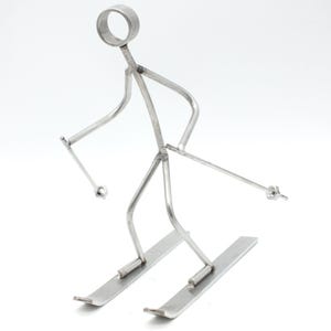 Könnte beinhalten: Eine silberfarbene Metallskulptur eines Skifahrers, gefertigt aus geschweißten Stäben und Metallteilen. Die Figur hat einen kreisförmigen Kopf, angewinkelte Arme mit Skistöcken, die in Muttern enden, und steht auf zwei flachen Metallskiern.