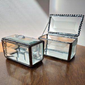Puede incluir: Dos joyeros rectangulares de cristal con tapas con bisagras. Una caja está cerrada y la otra está abierta, revelando el interior. Las cajas tienen un borde de metal plateado y una cadena decorativa. El cristal está facetado, creando un efecto de prisma.