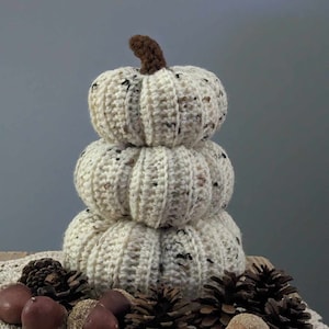 Peut inclure: Une pile de trois citrouilles crochetées de couleur crème avec des mouchetures brunes. Les citrouilles sont posées sur un napperon en crochet blanc avec des pommes de pin et des glands bruns.