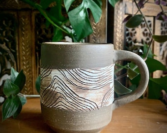 May include: A handmade ceramic mug with a brown base and rim, featuring a textured, carved design in a lighter brown hue. The mug has a rounded handle and a cylindrical shape, perfect for enjoying hot beverages.