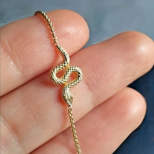 Puede incluir: Un brazalete de serpiente de oro con una cadena delicada. El encanto de la serpiente es detallado y tiene una superficie texturizada.