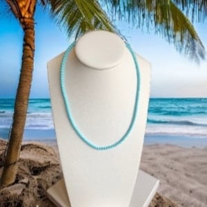 May include: A light blue beaded necklace on a white mannequin bust against a beach background.