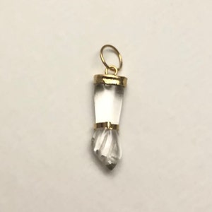 Könnte beinhalten: Ein Anhänger aus klarem Quarzkristall mit einer goldfarbenen Metalleinfassung. Der Kristall ist facettiert und hat eine spitze Spitze. Der Anhänger befindet sich auf einem weißen Hintergrund.