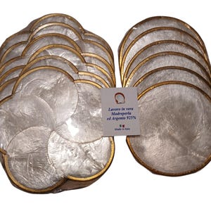 Op de afbeelding: Een set van 12 witte parelmoer-onderzetters met een gouden rand. De onderzetters zijn gevormd als een klavertje vier. De verpakking bevat een label met de tekst "Lavoro in vera Madreperla ed Argento 925% Made in Italy".