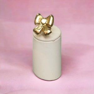 Può includere: Un anello dorato a forma di fiocco presentato su un espositore per anelli bianco. L'espositore è su una superficie rosa.