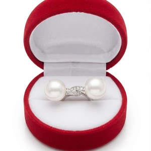 May include: A bypass ring featuring two white pearls and a silver-toned band with small clear stones set in a twisted crossover design, presented inside a round, red velvet-lined jewelry box with a white interior cushion.