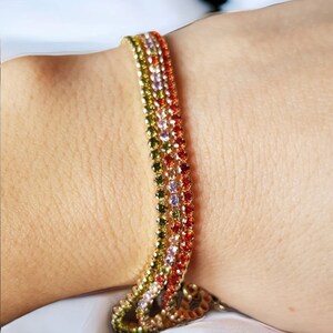 May include: A gold-toned bracelet with multiple rows of colorful gemstones. The gemstones are arranged in a gradient of colors, including green, yellow, orange, red, and purple. The bracelet is worn on a wrist.