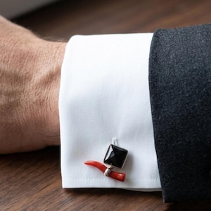 Puede incluir: Primer plano de un puño de camisa blanca con un gemelo negro cuadrado y un cuerno rojo. El gemelo es plateado y tiene un diseño de cuerno rojo.