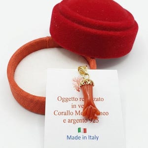 Puede incluir: Un colgante de color coral en forma de mano, con detalles dorados, cuelga frente a una caja de joyería de terciopelo rojo. Se ve una tarjeta blanca con texto italiano y una pequeña bandera italiana. El colgante está hecho de coral y plata.