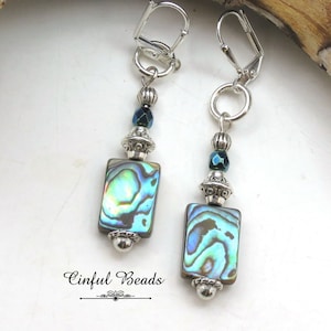 May include: A pair of silver earrings with abalone shell and blue glass beads. The earrings have a rectangular abalone shell bead, a silver bead, a blue glass bead, and a silver hook.
