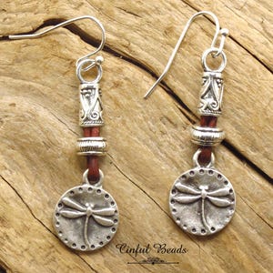 May include: Silver dragonfly earrings with brown leather cord and silver beads. The earrings have a silver hook closure.