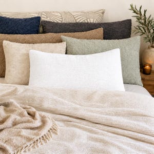 May include: A collection of rectangular throw pillows in various colours including white, beige, sage green, dark grey, and navy blue, featuring textured fabric surfaces. A matching beige throw blanket with fringe edges rests in the foreground.