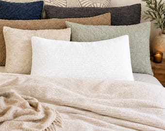 May include: A collection of rectangular throw pillows in various colors including white, beige, sage green, dark gray, and navy blue, featuring textured fabric surfaces. A matching beige throw blanket with fringe edges rests in the foreground.