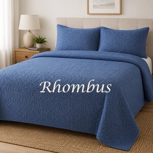 May include: A blue bedspread and two matching pillow shams featuring a textured, repeating geometric pattern. The word Rhombus is printed in white across the centre of the bedspread. The bedding set is displayed on a bed with a headboard.