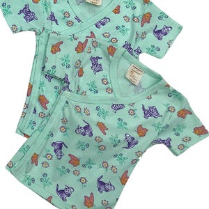 May include: Two turquoise baby onesies with short sleeves. The onesies have a wrap-style closure and feature a print of purple kittens, orange butterflies, and green leaves on a light turquoise background. The text "3 Months" is printed on the label of each onesie.
