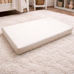 May include: A rectangular, off-white baby changing pad. The pad is placed on a plush, cream-colored rug. The changing pad is a solid color and appears to be made of a soft, durable material. The pad is designed for use in a nursery setting.