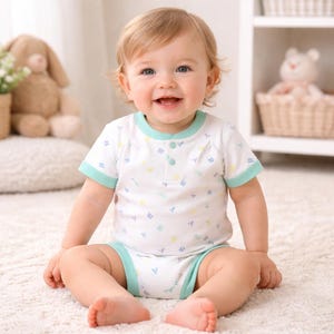 May include: A smiling baby wearing a white onesie with a light blue trim and a pattern of colorful letters. The onesie has short sleeves and a button-up front. The baby is sitting on a white rug.