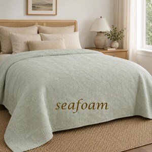 May include: A seafoam green bedspread with a textured, geometric pattern covering the surface. The word "seafoam" is printed in brown text on the lower centre of the fabric. The bedspread is draped over a bed with several beige pillows.