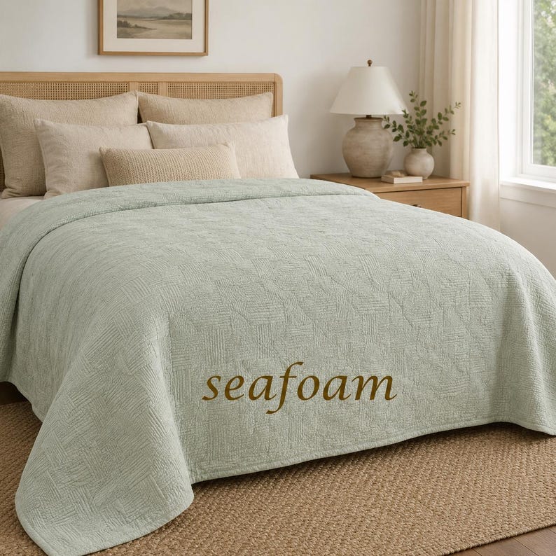 May include: A seafoam green bedspread with a textured, geometric pattern covering the surface. The word "seafoam" is printed in brown text on the lower centre of the fabric. The bedspread is draped over a bed with several beige pillows.