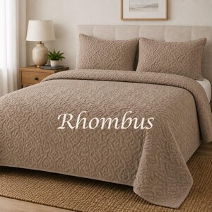 May include: A light brown bedspread and two matching pillow shams featuring a raised, textured geometric pattern. The word Rhombus is printed in white across the centre of the bedspread. The bedding set is displayed on a bed with a headboard.
