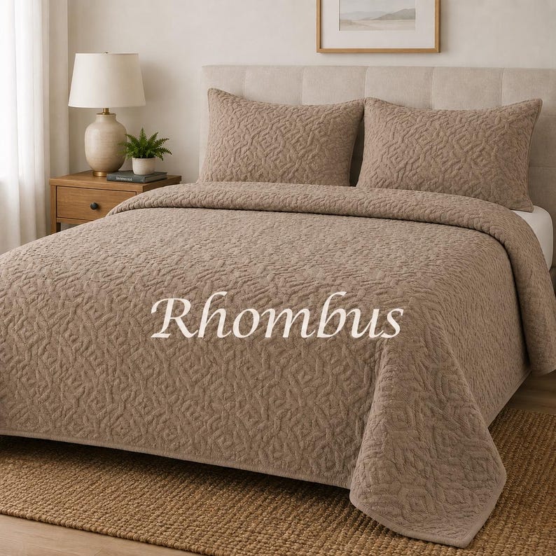 May include: A light brown bedspread and two matching pillow shams featuring a raised, textured geometric pattern. The word Rhombus is printed in white across the centre of the bedspread. The bedding set is displayed on a bed with a headboard.