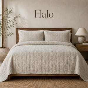 May include: A beige textured quilt set on a wooden bed frame with two matching pillows. The word Halo is printed in dark brown letters on the wall above the bed. The bedding features a subtle grid pattern and a soft, woven appearance.