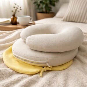 May include: A U-shaped cream-colored neck pillow with a textured fabric surface rests on top of a round, flat, cream-colored cushion. A bright yellow circular fabric base with a drawstring tie is positioned underneath the round cushion.